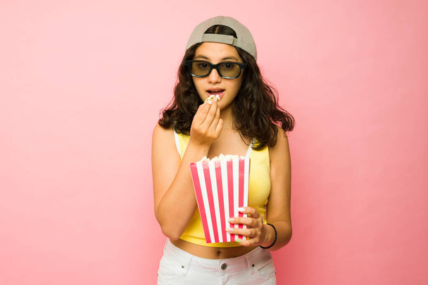 Beautiful thirteen-year-old teen girl eating popcorn while watching a 3d movie at the cinema against a pink background - Photo, Image