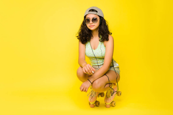 Ισπανόφωνη έφηβη κοπέλα που διασκεδάζει πατινάζ χρησιμοποιώντας παλιά πατίνια και καλοκαιρινά γυαλιά ηλίου δίπλα στο κίτρινο αντίγραφο χώρο - Φωτογραφία, εικόνα
