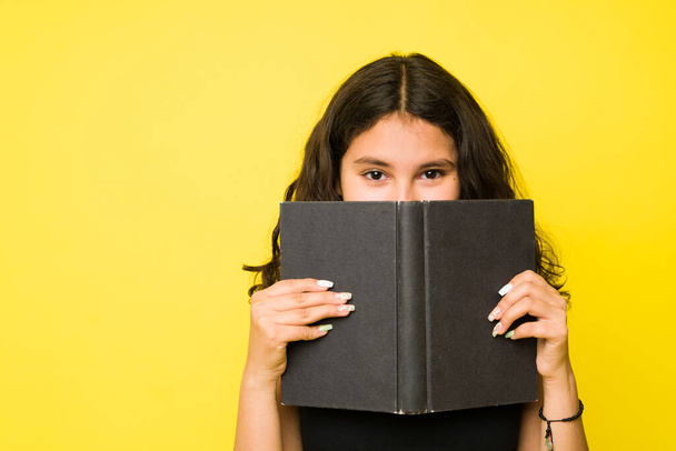 Schattig verlegen tiener meisje verbergen achter een boek tijdens het lezen en genieten van een roman tegen een gele studio achtergrond - Foto, afbeelding