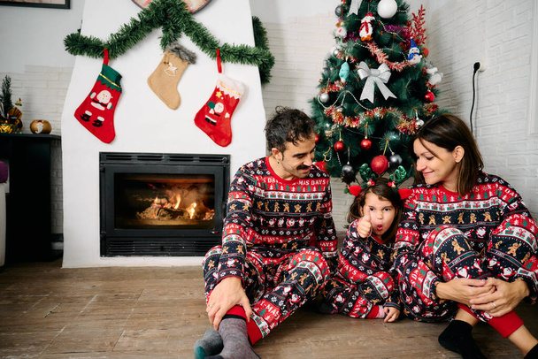 a family, with the father, mother, and daughter, wearing matching Christmas pajamas, taking family photos in front of the fireplace and a Christmas tree. The scene represents the joy, love, and togetherness of the holiday season - Фото, зображення