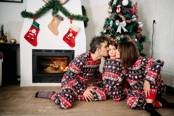 egy család, az apával, anyával és a lányukkal, ugyanolyan karácsonyi pizsamában, családi fotókat készítve a kandalló és egy karácsonyfa előtt. A jelenet az ünnepi szezon örömét, szeretetét és összetartását jelképezi. - Fotó, kép