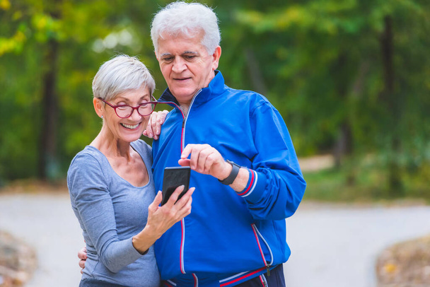 Smiling senior active couple walking in park looking at smartphone - Photo, Image