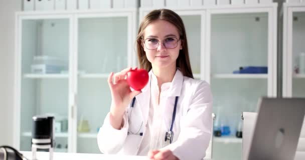 Professional woman in glasses holds heart toy with smiling and delighted expression. Female specialist enjoys working doctor slow motion - Footage, Video