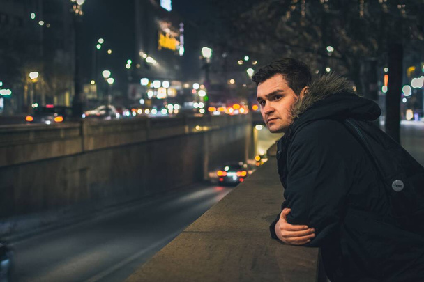 A young caucasian man leaning on a low stone wall on the street at night - Φωτογραφία, εικόνα