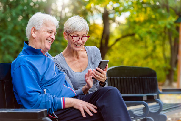 Uśmiechnięta aktywna para siedzi na ławce i patrzy na smartfona - Zdjęcie, obraz