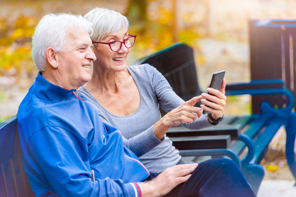 Улыбающаяся старшая активная пара сидит на скамейке и смотрит на смартфон - Фото, изображение
