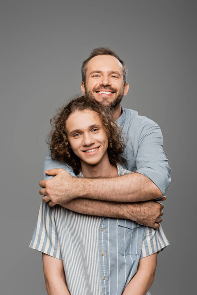 cheerful man hugging curly teenager son and looking at camera isolated on grey  - Foto, imagen