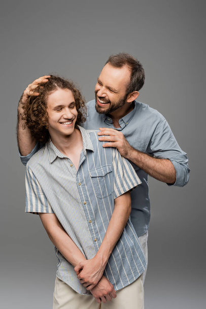 cheerful dad touching curly hair of his teenager son and smiling isolated on grey  - Foto, Imagen