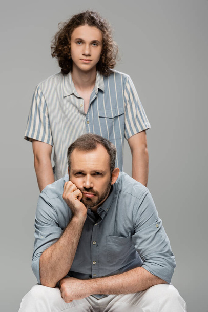 bored father looking at camera next to curly teenage son isolated on grey  - Фото, зображення