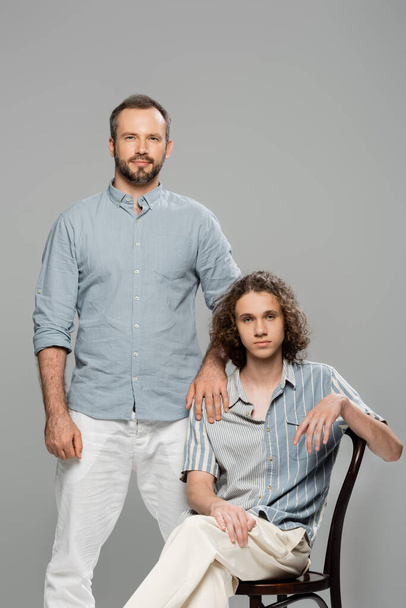 good looking dad standing next to teenage son with curly hair isolated on grey  - Фото, изображение