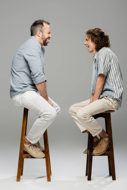 side view of funny dad and teenage son grimacing at each other while sitting on high chairs on grey  - Zdjęcie, obraz