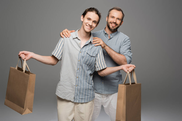 feliz adolescente sosteniendo bolsas de compras cerca alegre papá en gris  - Foto, imagen