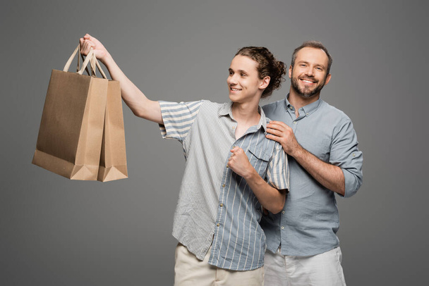 happy teenage boy holding shopping bags near positive father isolated on grey  - 写真・画像