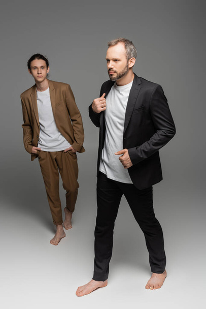 full length of well dressed man walking with bare feet next to stylish teenage boy on grey  - Fotografie, Obrázek