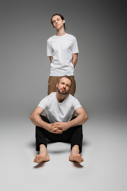 plná délka vousatého otce a dospívajícího syna v bílých tričkách pózujících na šedi  - Fotografie, Obrázek