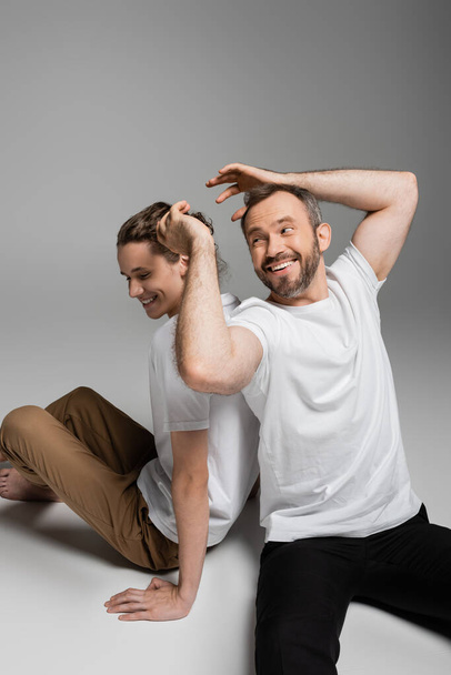 cheerful father having fun with teenage son in white t-shirt and sitting on grey  - Фото, изображение