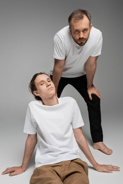 bearded father and teenage son in white t-shirts posing while looking at camera on grey  - Фото, изображение