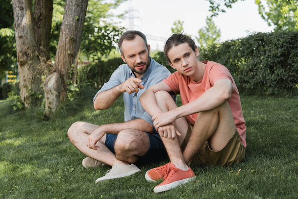 szakállas férfi, aki tinédzser fia felé mutat, miközben együtt ülnek a zöld pázsiton.  - Fotó, kép