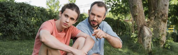 bearded man pointing away near teenager son while sitting together in park, banner  - Фото, изображение
