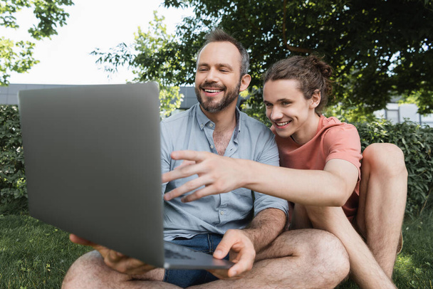 souriant père en utilisant un ordinateur portable à côté de garçon adolescent heureux tout en étant assis ensemble sur la pelouse verte  - Photo, image