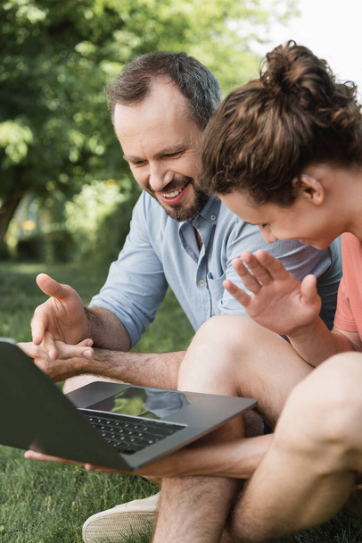 happy father pointing at laptop next to teenage boy while sitting together on green lawn  - Фото, зображення