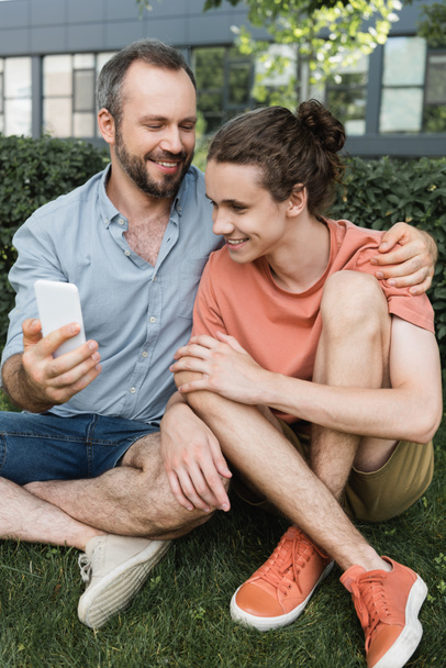 happy man holding smartphone near teenager son while sitting together in green park  - Photo, Image