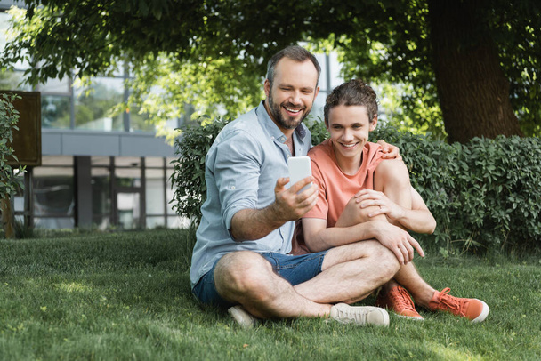 šťastný muž brát selfie s teenager syn zatímco sedí spolu v zeleném parku  - Fotografie, Obrázek