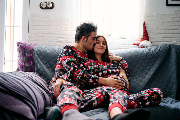 una joven pareja heterosexual, vestida con pijamas de Navidad, compartiendo un beso amoroso en el sofá de su casa. La escena captura el calor y la comodidad de la temporada navideña, llena de amor, romance y unión. - Foto, imagen