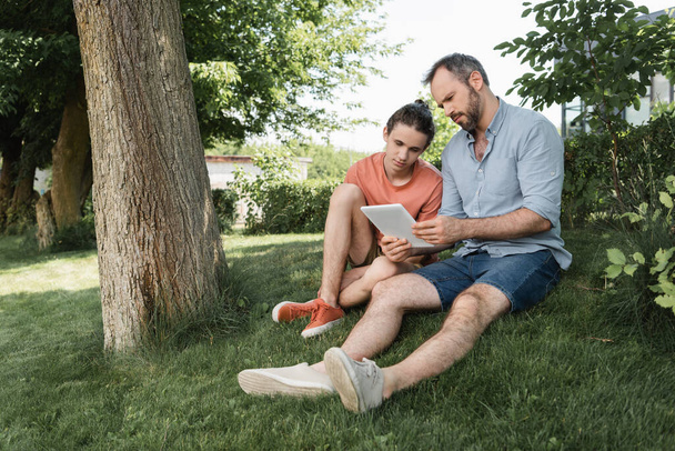 бородатый отец и сын-подросток смотрит на цифровой планшет, сидя рядом с деревом в парке  - Фото, изображение