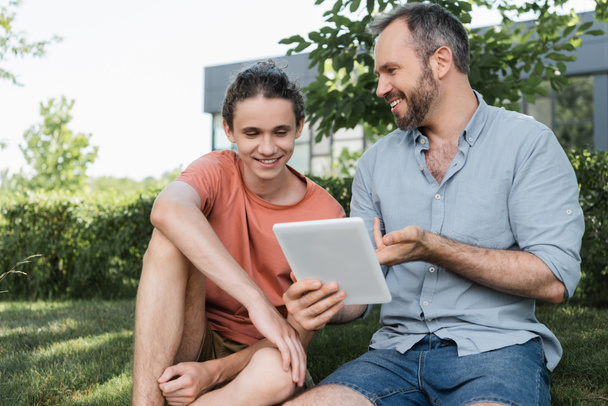 bearded dad and happy teenager son looking at digital tablet while sitting in green park  - 写真・画像