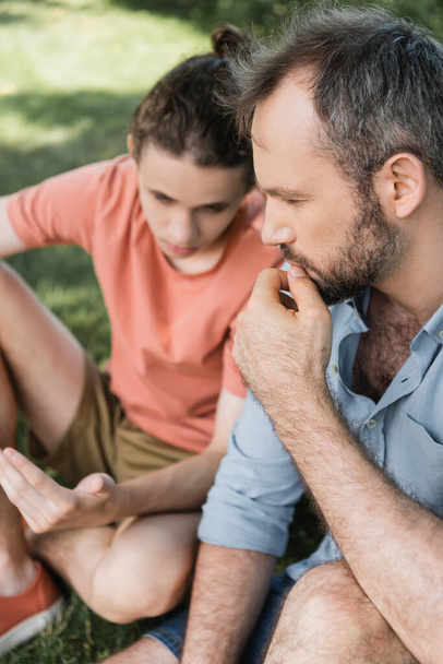 high angle view of pensive and bearded father sitting next to teenage son in green park  - Fotoğraf, Görsel