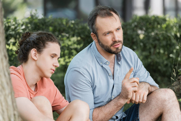 vousatý otec ukazuje pryč, zatímco sedí vedle dospívající syn a mluví v zeleném parku  - Fotografie, Obrázek