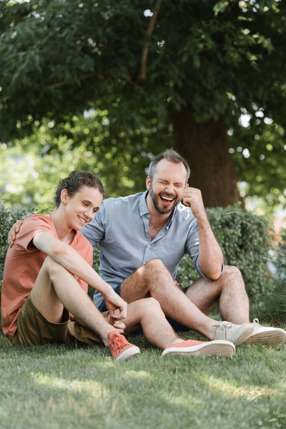 cheerful father and son sitting in summer clothes on green lawn in park  - Foto, Imagen