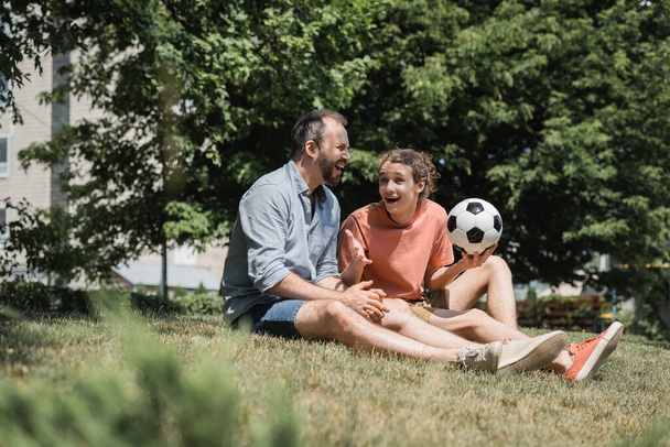 excited teenage boy sitting near father and soccer ball in green summer park  - Фото, изображение