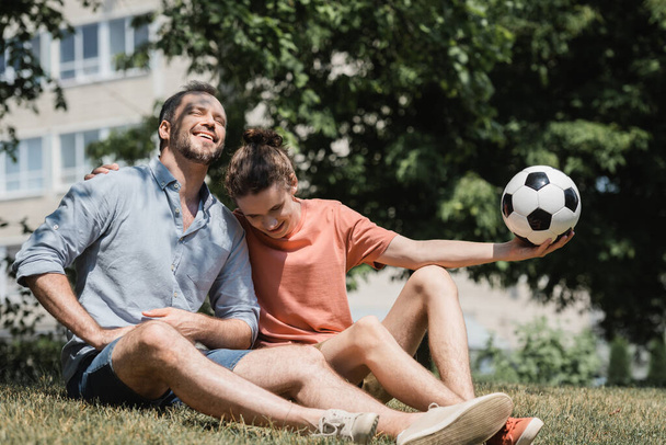 adolescente sentado perto de pai alegre e segurando bola de futebol no parque de verão verde  - Foto, Imagem