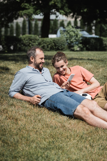 vousatý muž s úsměvem při použití smartphonu a odpočinku na trávníku v blízkosti dospívající syn po hraní fotbalu  - Fotografie, Obrázek