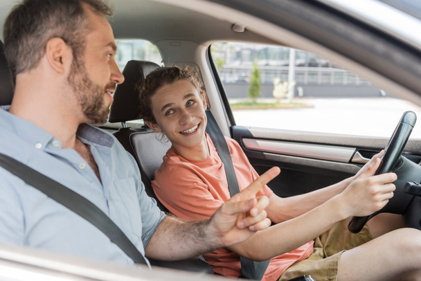 happy teenaged boy sitting next to dad while learning how to drive car  - Photo, Image