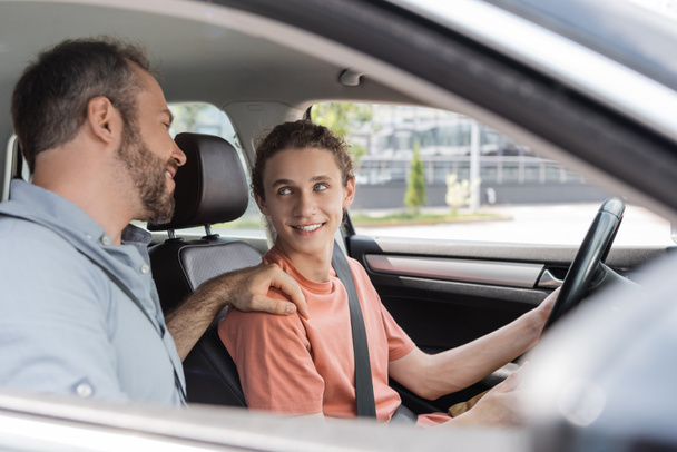 alegre padre poniendo la mano en el hombro de hijo adolescente mientras le enseña a conducir coche  - Foto, imagen