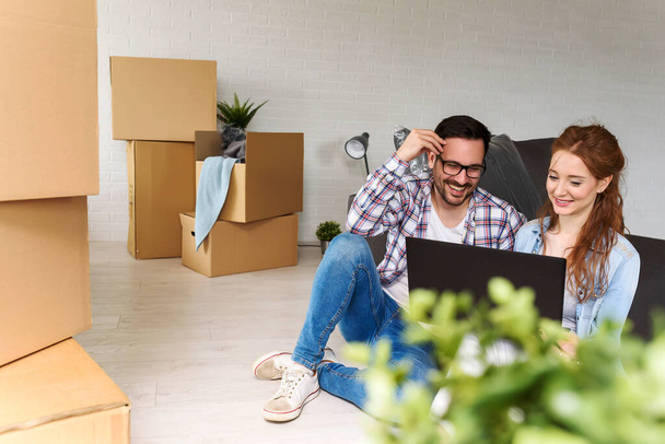 Beautiful young couple sitting among cardboard boxes in their new apartment making plans with laptop - 写真・画像