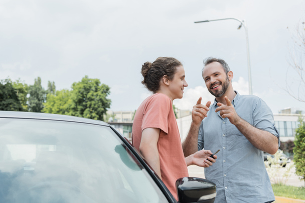 boldog szakállas apa ujjal mutogató közel tizenéves fiú áll autó mellett kulcsokkal  - Fotó, kép