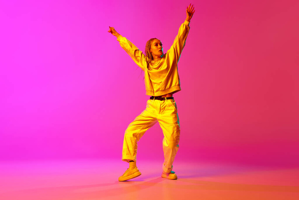 Portrait with one young girl, dancer with dreadlocks dancing with hands up over gradient pink background in neon light. Contemporary dance style, motion, art, movement, inspiration, hobby, ad concept - Photo, image