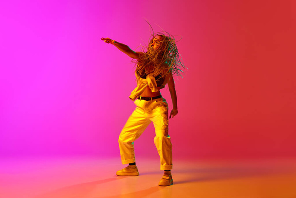 Ένας επαγγελματίας hip-hop χορευτής φορώντας ρούχα μόδας κινείται με έμπνευση πάνω από κλίση ροζ φόντο σε νέον φως. Έννοια του σύγχρονου χορού στυλ, κίνηση, χόμπι, τέχνη, διαφήμιση - Φωτογραφία, εικόνα