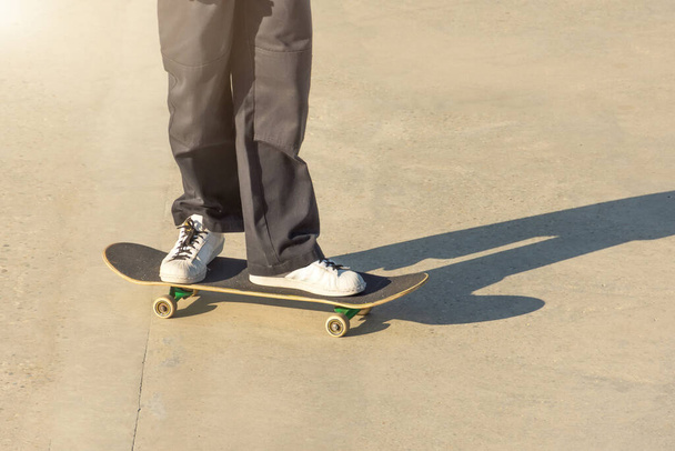 Хлопець в чорних вантажних джинсах і кросівках їде на бетонній підлозі зі скейтбордом, кладе на нього ногу
 - Фото, зображення