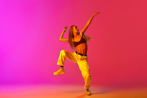 Portrait of young stylish woman, hip-hop dancer training in casual clothes over gradient pink background in neon light. Youth culture, movement, street style, fashion, action, contemporary dance style - Photo, Image