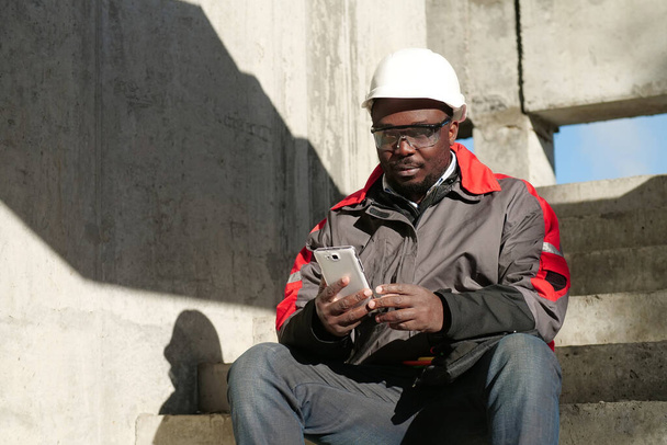 Afrikai amerikai munkás kemény kalapban és védőszemüvegben okostelefonnal ül az építkezésen. A munkás fehér okostelefont tart a kezében és a kamerába néz - Fotó, kép