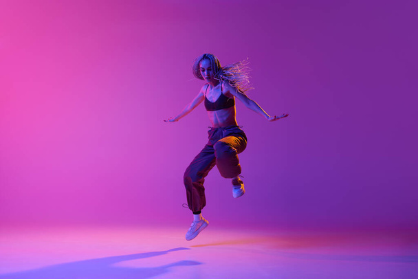 Импровизация. Одна танцовщица, женщина в обычной одежде, прыгающая в движении над градиентным фиолетовым неоновым фоном. Мбаппе современного стиля танца, вдохновения, движения, движения, моды, искусства - Фото, изображение