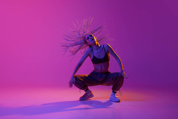 Twerk. Een jong charmant meisje, professionele danser met vliegend haar in casual kleding dansend over paarse studio achtergrond in neon licht. Concept van hedendaagse dans, kunst, sport, mode - Foto, afbeelding