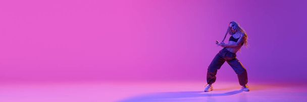 Wolny styl. Baner z atrakcyjną młodą kobietą, dziewczyną z warkoczami tańczącą solo z przyjemnością nad fioletowym tłem w neonowym świetle. Koncepcja tańca współczesnego, sztuki, sportu, mody, hobby - Zdjęcie, obraz