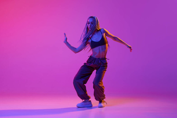 ヒップホップダンスバトル。ネオンの光の中で紫のスタジオの背景に感情を込めて踊るストリートスタイルの服を着た若い女の子。現代的なスタイル、動き、アート、動き、インスピレーション、広告の概念 - 写真・画像