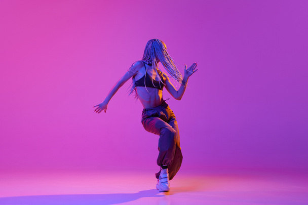 Szkolenie w pomieszczeniach. Portret młodej dziewczyny, tancerka z dredami tańcząca nad gradientowym fioletowym tłem w neonowym świetle. Koncepcja współczesnego stylu, ruchu, sztuki, ruchu, inspiracji, reklamy - Zdjęcie, obraz